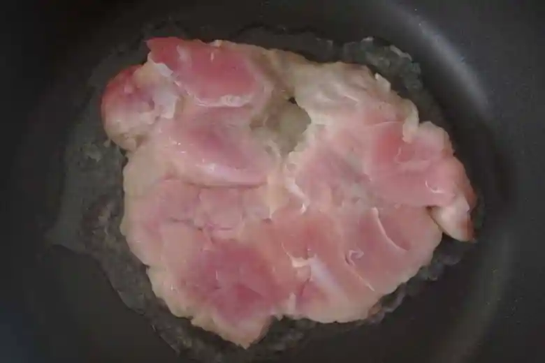 鶏もも肉を皮側を下にして黒いフライパンで焼いている写真です。肉がうっすらと白くなっています。油はひいていませんが、肉の周りに鶏自身の脂がしみでています。