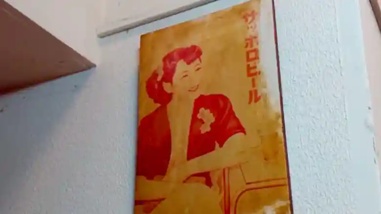「BEER & WINE GRILL」の壁に掲げられた古いポスターの写真です。胸と髪に花を飾った女性が描かれています。赤い文字でサッポロビールと書かれています。