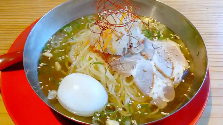 韓国式冷麺の写真です。金属製の銀色の丼に入っています。具材はキムチと大根なます、ゆで卵、糸唐辛子です。スープは透明で胡麻とネギが浮いています。