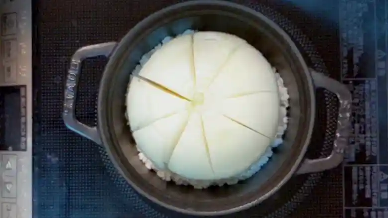 玉ねぎが入った鍋を上から撮影した写真です。米の上にまるごと１個の玉ねぎが置かれています。鍋は黒い鉄製で、直径が14cmです。玉ねぎには八等分に切込みが入っています。