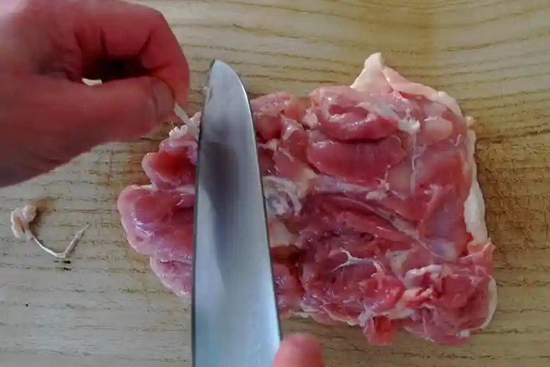 鶏もも肉の筋を包丁で切り取っている写真です。左手指で筋をひっぱりながら、右手に持った包丁で筋を切っています。