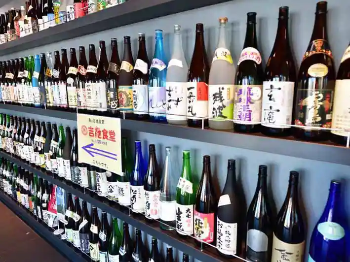 吉池食堂の入り口の壁に並ぶ一升瓶の写真です。壁一面、4段の棚に日本酒の一升瓶が並べられています。