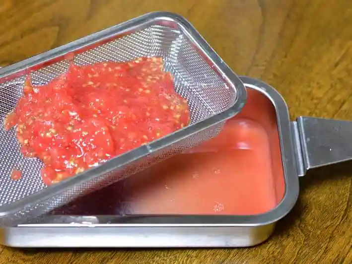 すりおろしたトマトの果肉を水切り網に入れて、水分を切っている写真です。トマトの果汁が受け箱にたまっています。