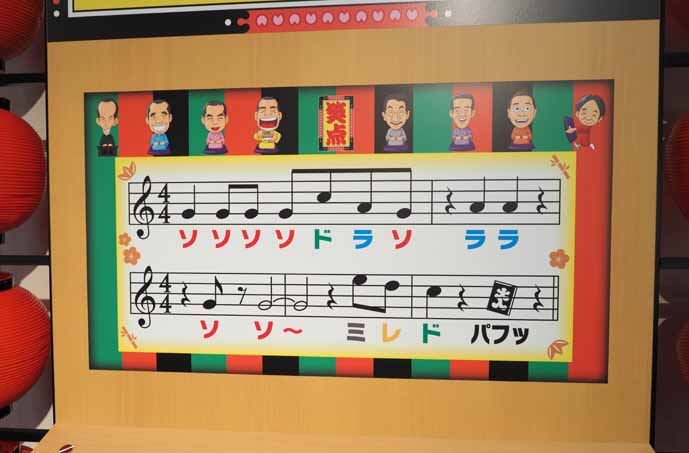 会場に設置されていた笑点のテーマ曲を奏でられるゲーム機の写真です。正面のボードにテーマ曲の楽譜が描かれています。音符には色がついています。手前には色のついたボタンがあります。ボタンを押すと音が出ます。音符と同じ色のボタンを押していくと笑点のテーマ曲を演奏できます。黒いボタンを押すと最後の「パフッ」という音が出ます。