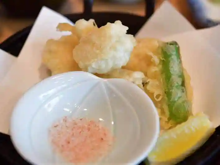 はもの天ぷらの写真です。黒い皿にシシトウといんげんの天ぷらも盛られています。梅塩とレモンが添えられています。