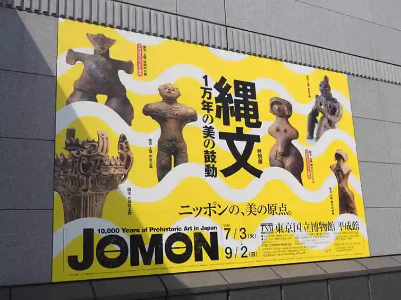 特別展「縄文－1万年の美の鼓動」の看板の写真です。東京国立博物館の平成館の入り口に設置されています。高さは3m、幅が4mほどの大きな看板です。背景は黄色で、国宝に指定されている土偶5体と火焔型土器1点が並んでいます。