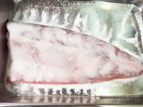 鯖を塩でしめている写真です。身が見えなくなるまで塩が振りかけられています。水分を下へ流すためパッドは斜めに設置しています。