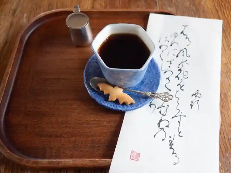 ホットコーヒーの写真です。コーヒーにはこうもりの形をしたビスケットが添えられています。コーヒーにも女将が筆でしたためた和歌の短冊がそえられています。「見渡せば柳桜をこき混ぜて都ぞ春の錦なりける」と墨で書かれています。