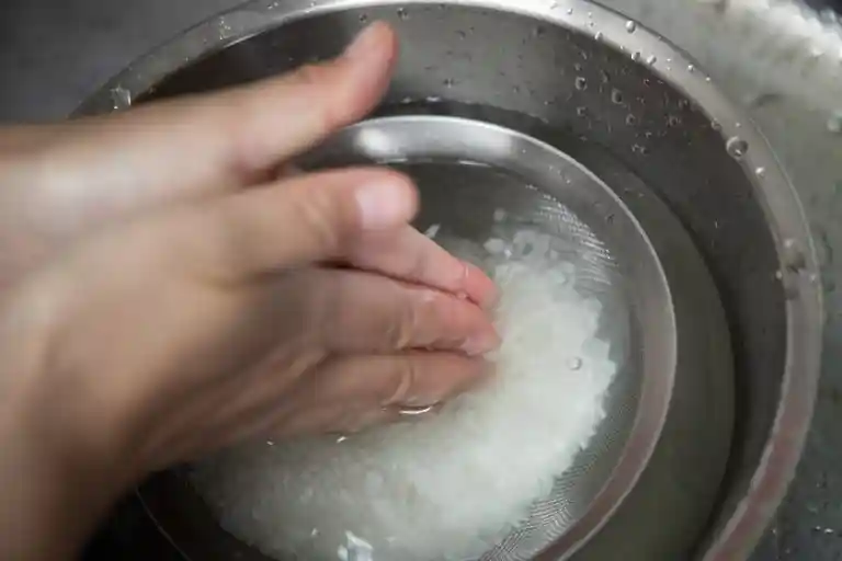 お米を洗っている写真です。大きなボールに水をくみ、ひとまわり小さいザルに米を入れて水につけています。手のひらで米を擦りつけるようにして洗っています。