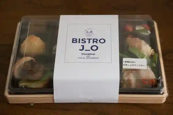 「BISTRO J_O 厳選欲張り重」が入ったプラスチック容器の写真です。縦20cm、横25cmの大きさです。蓋は透明で、白い紙で覆われています。紙には紫色の文字で「BISTRO J_O」と印刷されています。