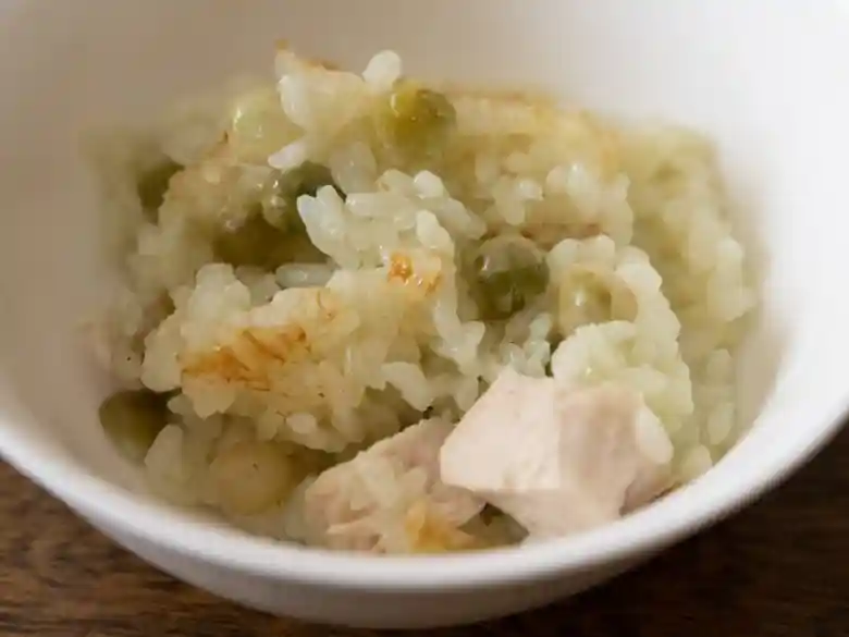 お茶碗によそった「スパイシ～青豆☆釜飯」の写真です。白いお茶碗にご飯がよそられています。焦げたご飯の上に緑色のグリーンピースとピンク色の鶏肉がのっています。
