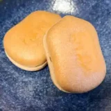 The photo shows Kuuya Monaka served on a blue round plate. It measures 5 cm in length and 3 cm in width and is shaped like a gourd. The word "空也（Kuuya）" is inscribed in Japanese in the center of the burnt glutinous rice skin. When the monaka is split in half, it is filled with crushed sweet bean paste.