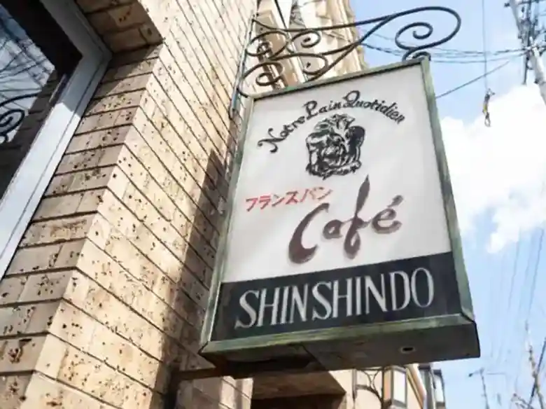 The photo shows a sign posted at the storefront of the cafe "Shinshindo" in front of Kyoto University's North Gate. The words "Notre Pain Quotidien" and "café" are written in French, and "French bread" is written in Japanese. Notre Pain Quotidien" means "our daily bread" in French.