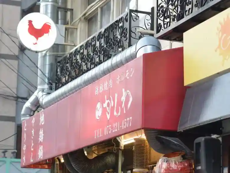 木屋町の「かしわや」の店先の写真です。白色の丸い看板に赤色でニワトリが描かれています。