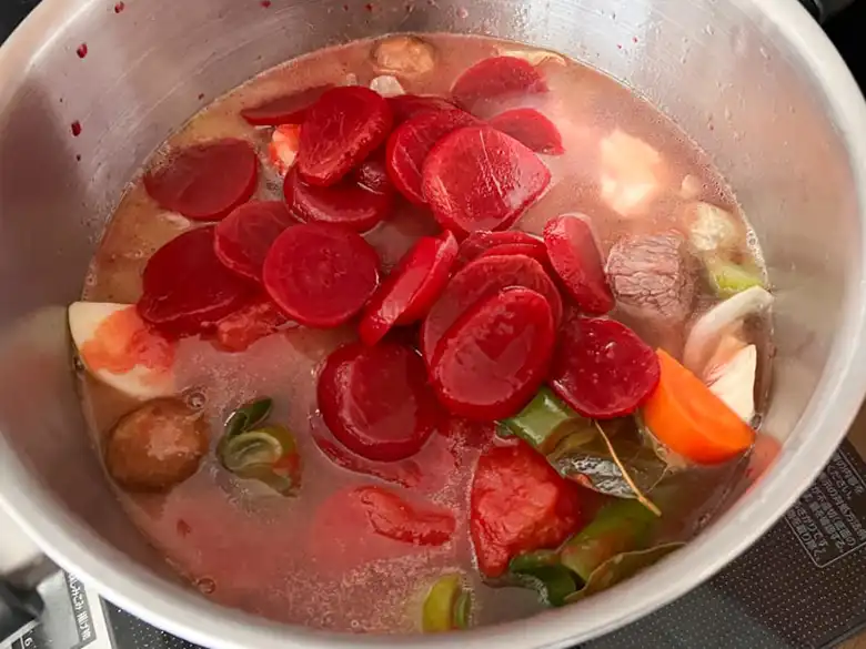 牛肉と野菜を炒めている鍋に、スープとビーツ、ビーツの汁を加えた写真です。スープが鮮やかな赤色になっています。