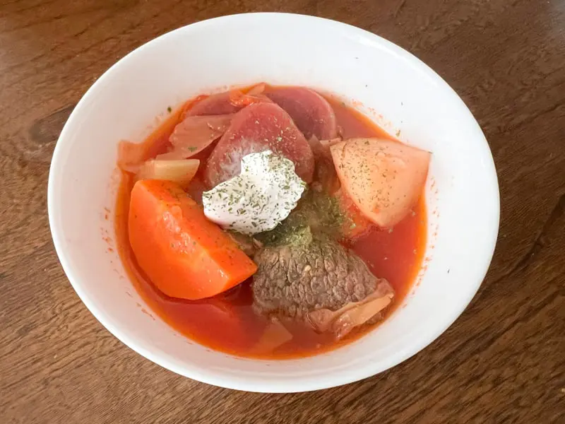 白い皿によそったボルシチにサワークリームを添えた写真です。赤いスープの中にビーツとキャベツ、じゃがいも、にんじんが見えます。