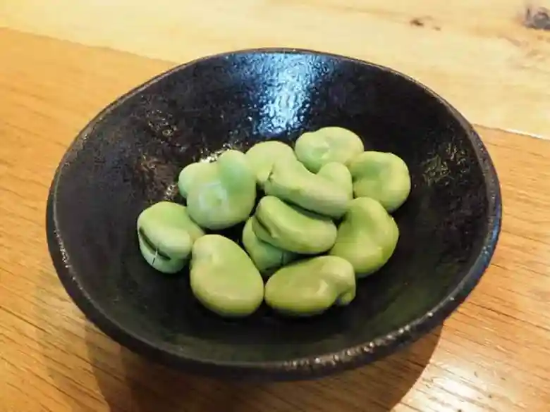 塩ゆでしたそら豆の画像です。黒い皿に緑色のそら豆が盛られています。そら豆のお歯黒の部分に切れ目が入っているので皮をむきやすくなっています。