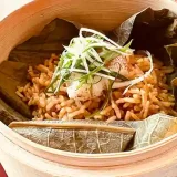 This photo shows Fried Rice with Steamed Dongxing Spots in XO Lotus Leaf Flavored Sauce. Fried rice wrapped in lotus leaves is served in a steamer basket. The fried rice is topped with steamed Dongxing Spots and chopped green onions.