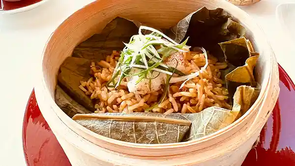 This photo shows Fried Rice with Steamed Dongxing Spots in XO Lotus Leaf Flavored Sauce. Fried rice wrapped in lotus leaves is served in a steamer basket. The fried rice is topped with steamed Dongxing Spots and chopped green onions.