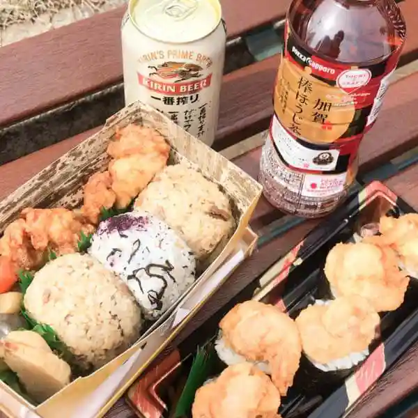 This picture shows a boxed lunch purchased at a JR Ueno Station kiosk. The lunch includes Onigiri (rice ball), fried chicken, and a beer, all placed on a bench.
