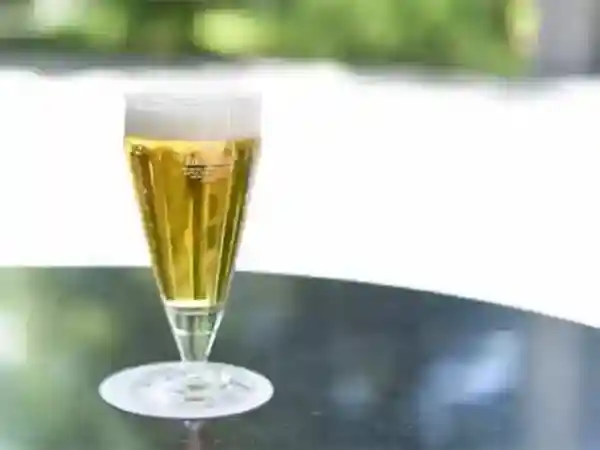 This photo shows a glass of beer poured on an outdoor table. The trees of the museum can be seen in the background behind the glass.
