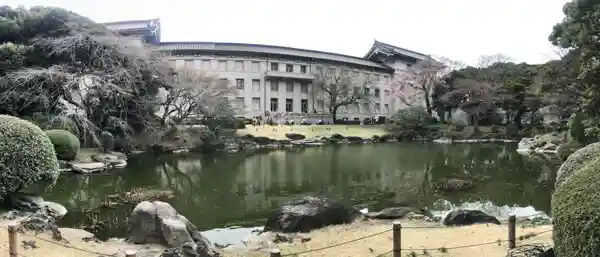 This photo shows the Honkan of the Tokyo National Museum as viewed from Rokusoan. Across the pond in the foreground is a grassy area in front of the Honkan. Cherry trees are planted around the pond.