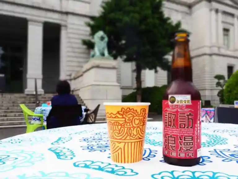 クラフトビールと「火焰型土器紙コップ」が表慶館前の野外テーブルの上に置かれた写真です。ビールは長野の諏訪浪漫です。