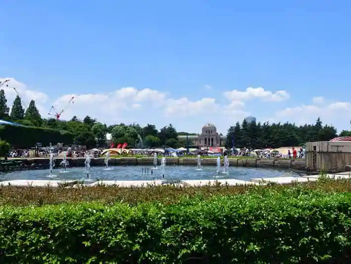 神宮外苑の噴水の写真です。噴水の奥に軟式野球場と絵画館が見えます。とても良い天気で青空に白い雲が浮かんでいます。