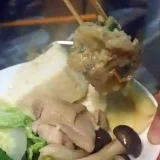 火が通った鶏団子を箸でつまんでいる写真です。鶏団子の直径は3cmぐらいです。小皿には鶏もも肉と白菜、もめん豆腐、しめじ、ニラが盛られています。