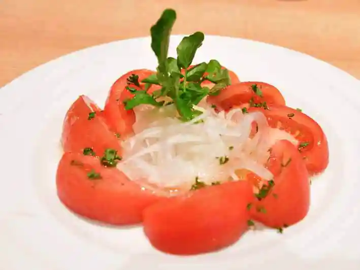 トマトサラダの写真です。白いお皿に8切れのトマトがリング状に並べられています。中央には玉ねぎと香草が添えられています。