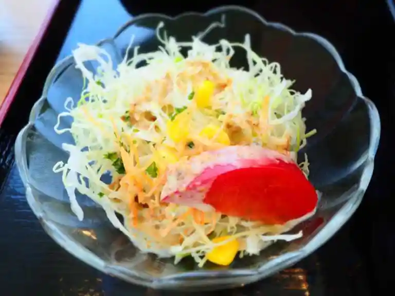 サラダの写真です。秋刀魚定食についてきました。透明なガラスの器の中に、刻まれたキャベツとにんじん、パセリ、コーン、トマトが盛られています。