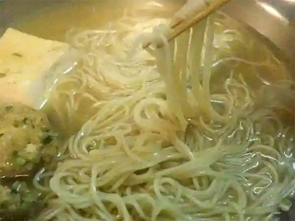 鍋からラーメンを箸でつまんでいる写真です。銀色の鍋の中には豆腐と鶏団子が残っています。