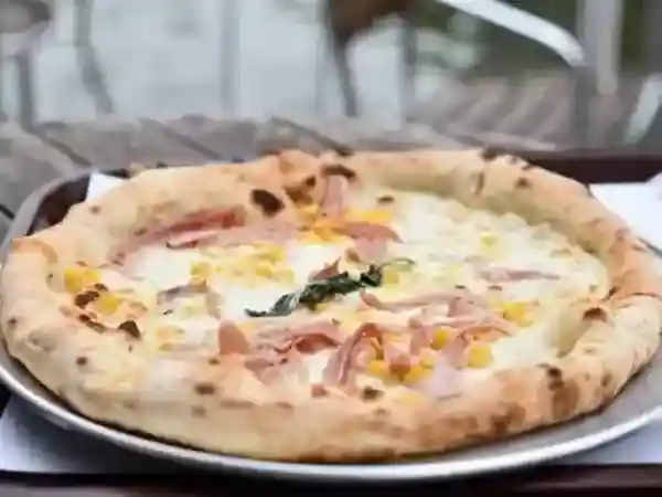 This photo displays a Neapolitan-style oven-baked pizza topped with prosciutto, corn, and rocket.