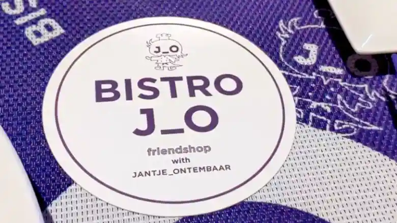 「BISTRO J_O」のコースターの写真です。円形で白色のコースターの上方にはジョーくんという鳥のキャラクターが紫色で描かれています。その下に「「BISTRO J_O」と紫色の文字で印刷されています。