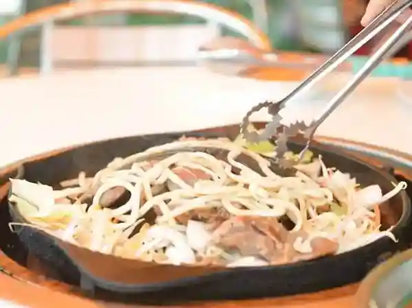 This image depicts the cooking of udon noodles in a Genghis Khan pot. The noodles are stir-fried and combined with cooked meat and vegetables to create a delicious meal