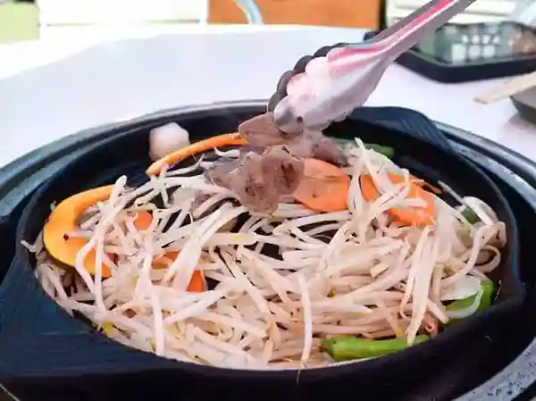 This photo shows lamb and vegetables cooking in an iron flat pot. The vegetables included in the dish are bean sprouts, carrots, and shishito peppers.