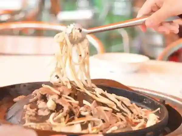 This photograph depicts the completed cooking of "Hokkaido Style Seasoned Lamb and Fried Udon Noodles." In the image, the udon noodles are picked up and lifted using tongs.