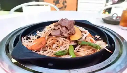 This photograph shows a dish featuring a raw lamb shoulder and assorted vegetables. The lamb is stir-fried with bean sprouts, carrots, and green peppers in a black iron pan.
