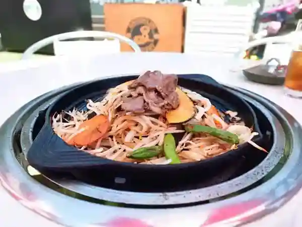 This photograph shows a dish featuring a raw lamb shoulder and assorted vegetables. The lamb is stir-fried with bean sprouts, carrots, and green peppers in a black iron pan.