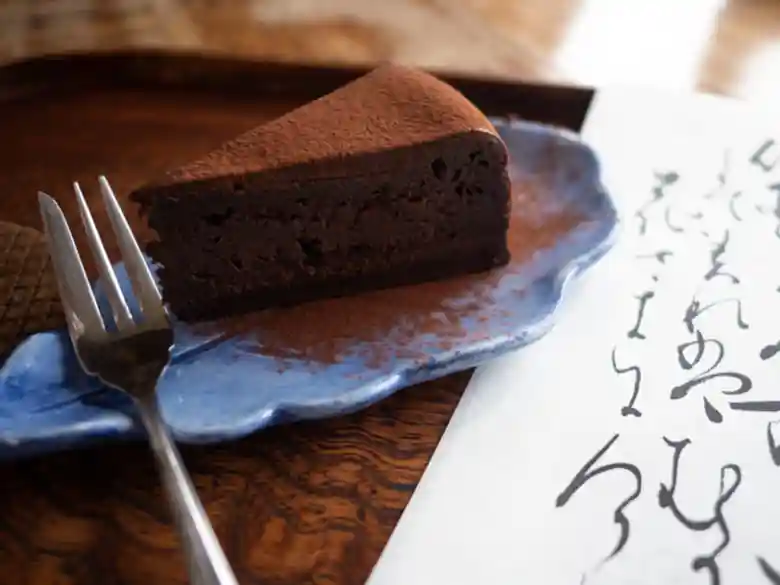 This photo shows a chocolate cake. It is served on a blue plate in the shape of a tree leaf. A strip of paper with a waka poem on it accompanies the cake. The proprietress of Yoshida Sanso wrote this poem in ink using a brush.