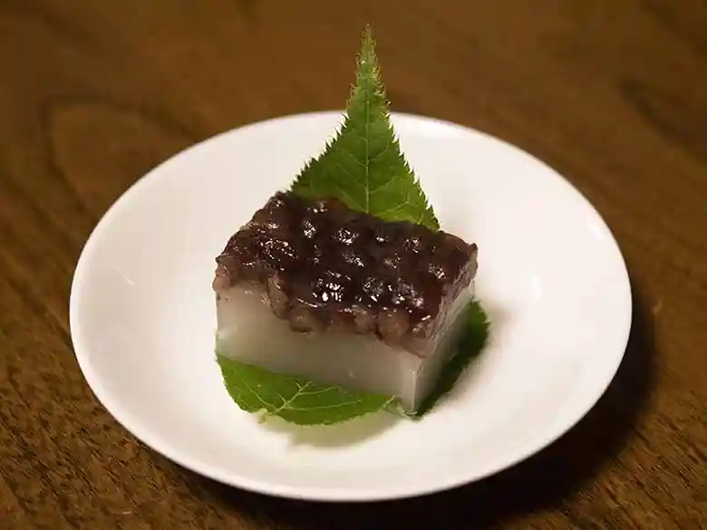 和菓子「水無月」の写真です。白い小皿に敷いた桜の葉に包まれています。白色のういろうの上に甘く煮た小豆がのっています。京都の水無月は三角形ですが、東京で購入したこの水無月は四角形でした。理由は分かりません。