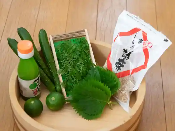 寿司桶に入ったすし飯のかやくの写真です。左から丸ごとのすだち、すだち酢の瓶、きゅうり、木の芽、青じそ、袋に入った白ごまが並べられています。