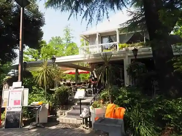 This image depicts the Hibiyasaro, with outdoor tables and chairs on the ground floor. The restaurant is situated amidst lush greenery, with numerous trees nearby.