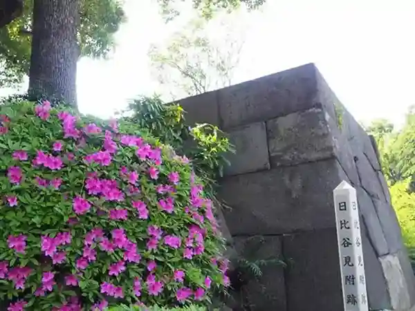 This photograph depicts the stone walls of Edo Castle, which still stand in Hibiya Park. A sign in front of the wall reads "Hibiya Mitsuke Site." In front of the stone wall, azalea flowers are in full bloom.