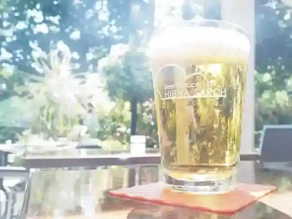 This photograph showcases draft beer poured into a glass mug on an outdoor table, with the trees of Hibiya Park in the background.