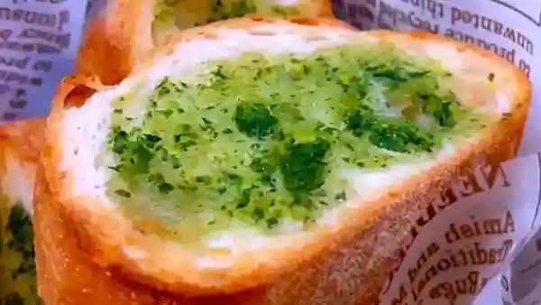 This picture shows garlic toast wrapped in paper with the French language printed. The surface is flavored with basil and garlic oil and has a green tint.