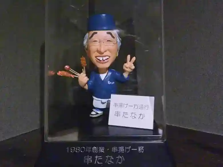 This photo shows a doll modeled after the master of Kushi-Tanaka. The doll is about 15 cm tall. The master is laughing and holding three Kushiage in his right hand and making a V-sign with his left hand. A signboard reading "Kushiage One Way Kushi-Tanaka" stands beside the doll.