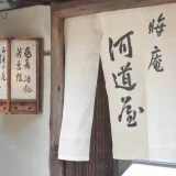 This photo shows the front of a soba noodle shop called Misoka-an Kawamichi-ya. A white "Noren" curtain hangs at the entrance. The name "Misoka-an Kawamichi-ya（晦庵 河道屋）" is written on the Noren in black letters.