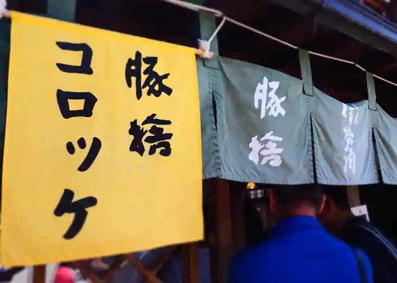 This picture shows the shop curtain of Butasute. The words "Butasute" and "Korokke" are written in black letters on the yellow fabric in Japanese.