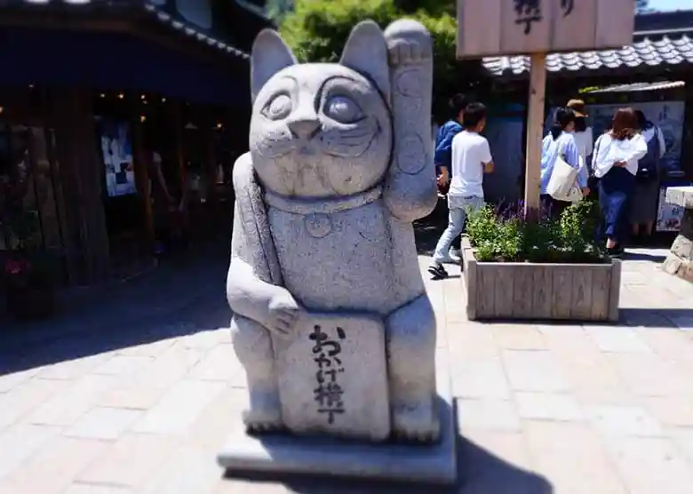 This picture shows a beckoning cat ornament placed at the entrance of Okage Yokocho. It is about 1.5m high. The beckoning cat is raising its left hand in greeting.